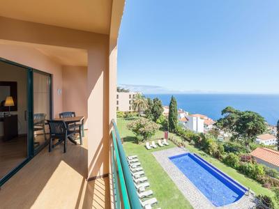 Balkon mit Stühlen und Meerblick, daneben ein Pool und Gartenbereich.