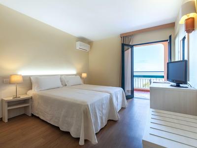 Helles Hotelzimmer mit Doppelbett, Nachttischlampen und Blick auf das Meer durch offene Balkontür.