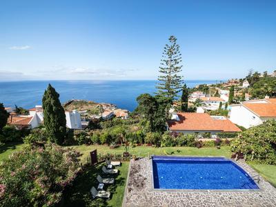 Blick auf einen blauen Pool und umliegende Häuser mit Meer im Hintergrund unter klarem Himmel.