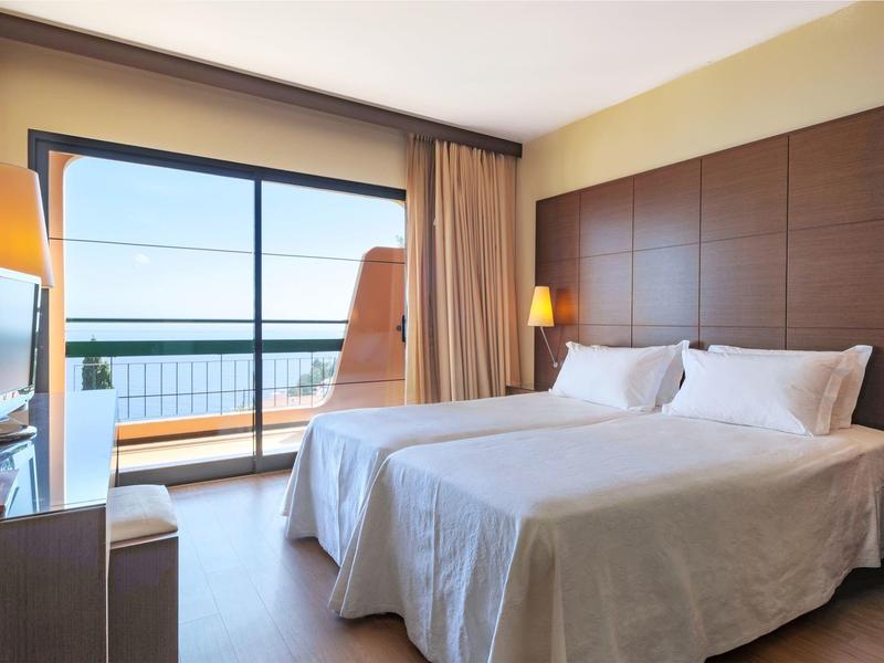 Hotel room with two beds, large window, and balcony overlooking the sea.