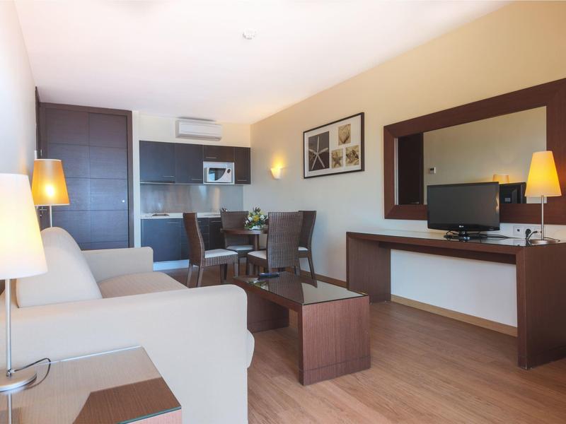 Modern hotel room with sofa, TV, table, and chairs on wooden floor.