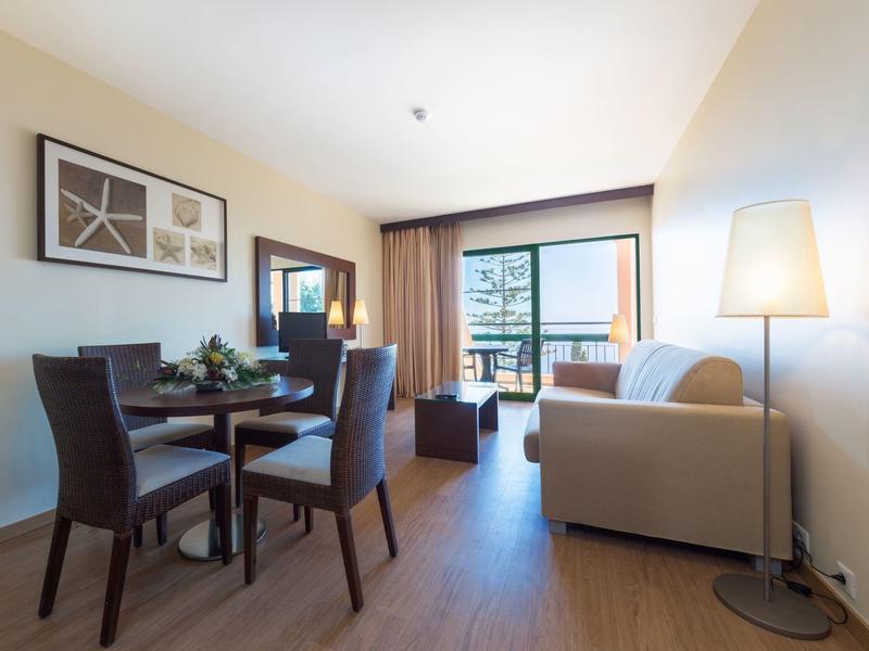 Modern hotel room with dining table, sofa, and balcony overlooking the sea.