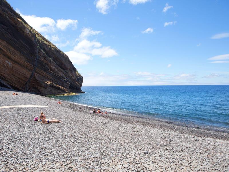 Pebble beach with few people, cliffs on the left, and calm sea under blue sky