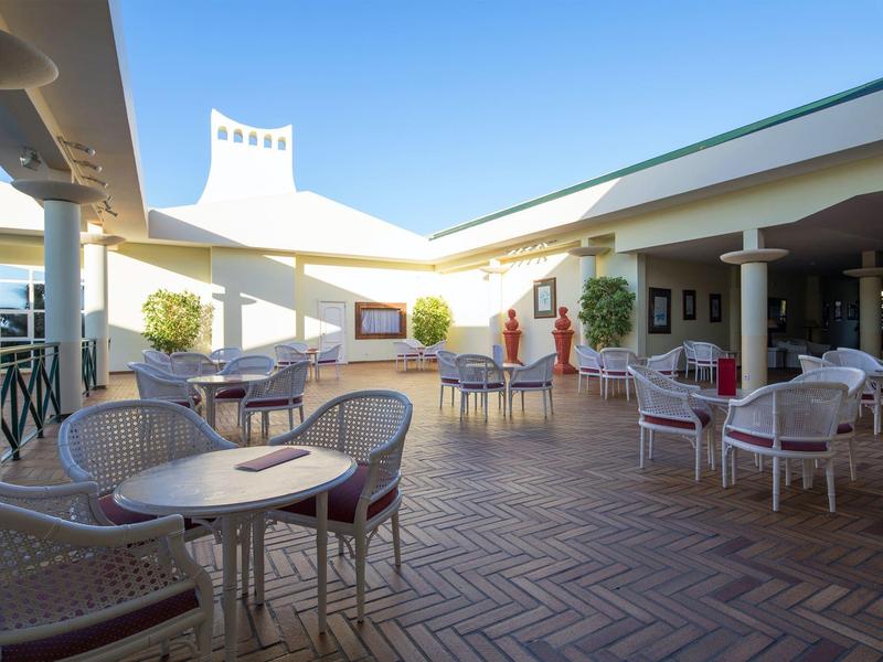 Bright outdoor terrace with multiple tables and chairs, sunshade, and blue sky.