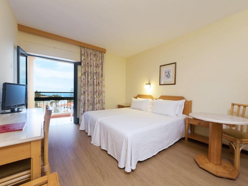 Brightly furnished hotel room with double bed, desk, TV, and balcony with view.