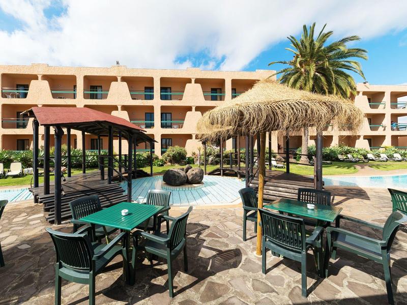 Hotel terrace with green chairs and tables, straw parasols, and palm trees in the background.