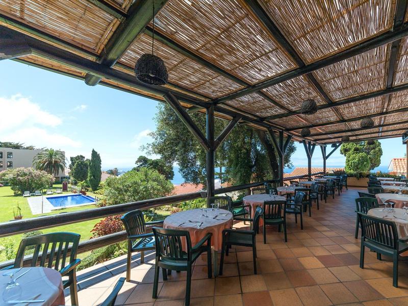 Covered terrace with tables and chairs overlooking garden with pool and sea in the background.
