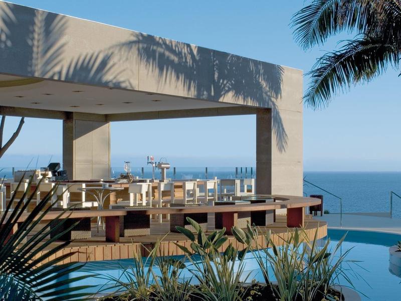 Offener, moderner Pavillon mit Holzmöbeln neben Swimmingpool und Meerblick bei klarem Himmel.