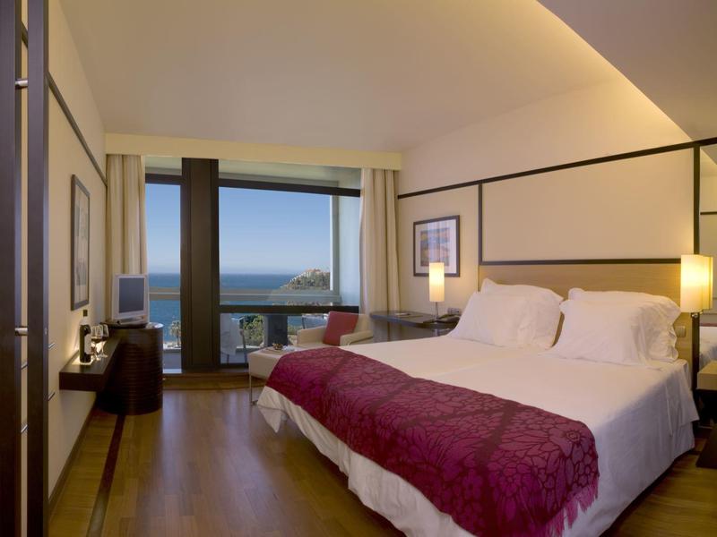 Modernes Hotelzimmer mit großem Bett, Holzboden, Balkon und Meerblick bei Dämmerung.