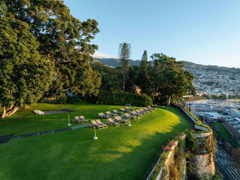 Grüner Hang mit Liegestühlen und Bäumen, im Hintergrund Berge und blauer Himmel.