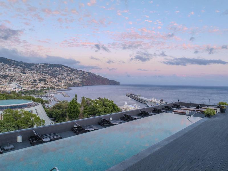 Infinity-Pool mit klarem Wasser, im Hintergrund Berg, Stadt und ruhiges Meer bei Abenddämmerung.
