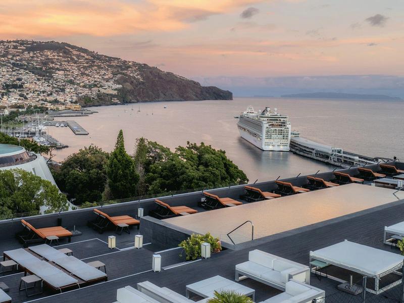 Blick auf Hafen mit Kreuzfahrtschiff, dahinter Hügel mit Häusern und farbigem Sonnenuntergangshimmel.