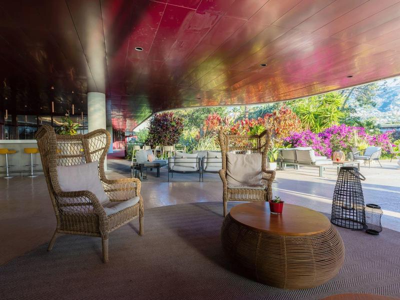 Offener Sitzbereich mit Rattanstühlen, Holztisch und blühenden Pflanzen vor Bergkulisse.