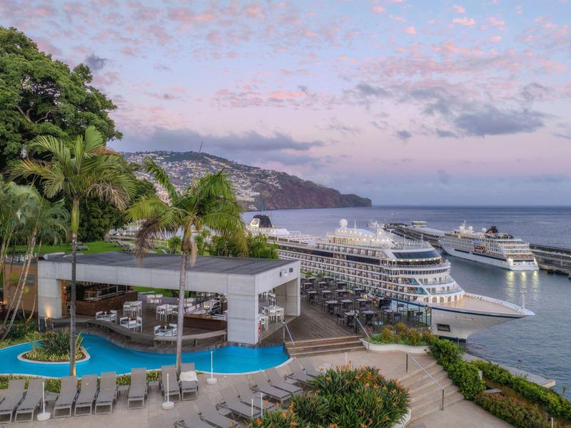 Kreuzfahrtschiffe am Hafen, neben Pool mit Liegestühlen und grünen Palmen bei Sonnenuntergang.