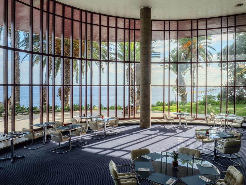 Großer, heller Raum mit bodentiefen Fenstern, modernen Stühlen und Blick auf Palmen und Meer.