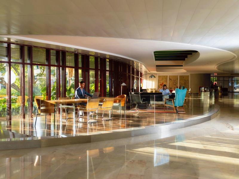 Großer, heller Innenraum mit glatten Böden, runden Tischen und großen Fenstern, Personen sitzen.