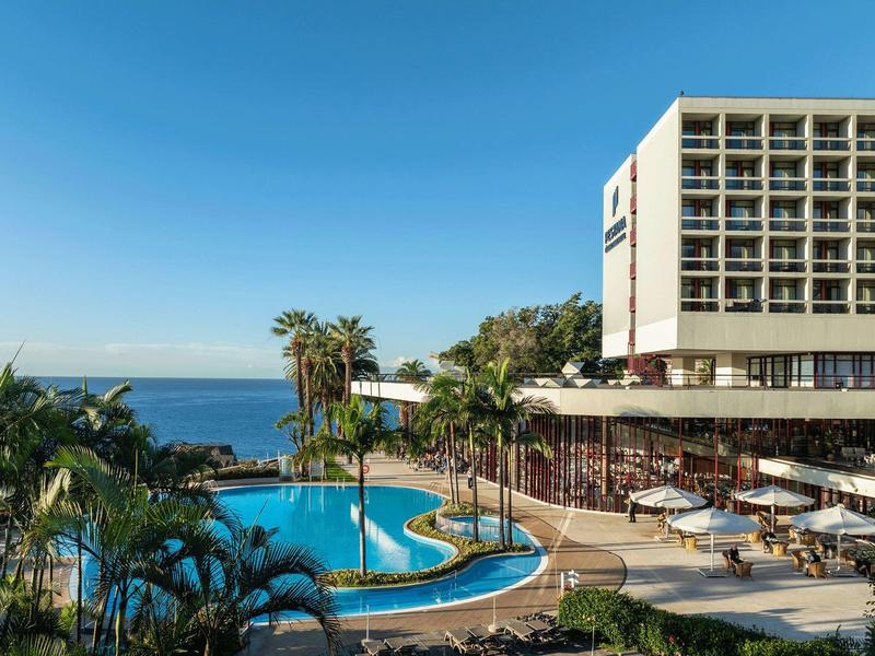 Hotel mit großem Pool, Palmen und Meerblick unter klarem blauen Himmel.