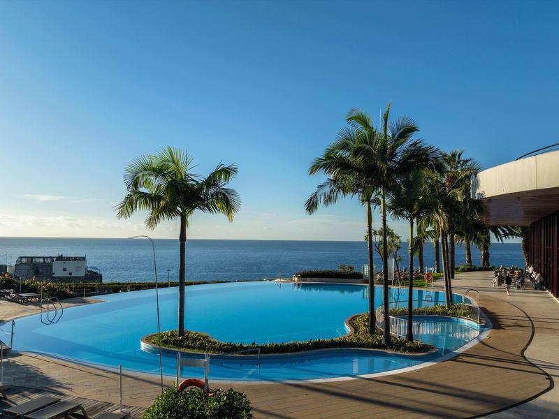 Infinity-Pool mit Palmen, Sonnenschirmen und Meerblick bei klarem Himmel am Tag.