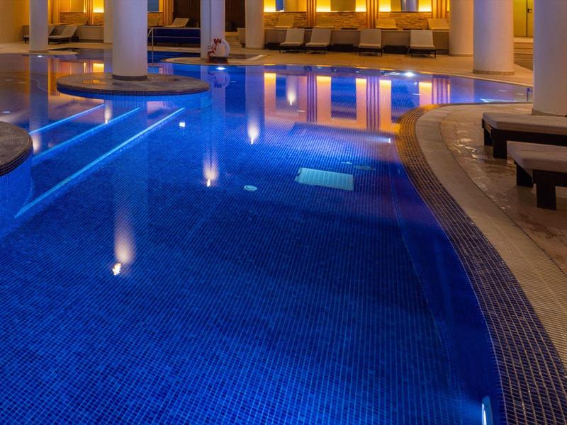 Innenpool mit blauen Kacheln, warmen Lichtern und komfortablen Liegen rundherum.