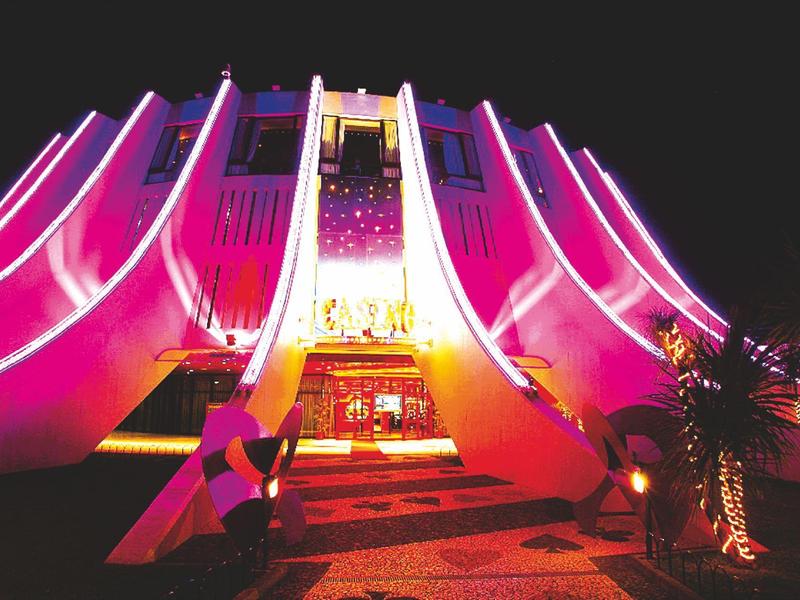 Modernes Gebäude mit wellenförmiger Fassade, beleuchtet in Pink und Lila, vor dunklem Nachthimmel.