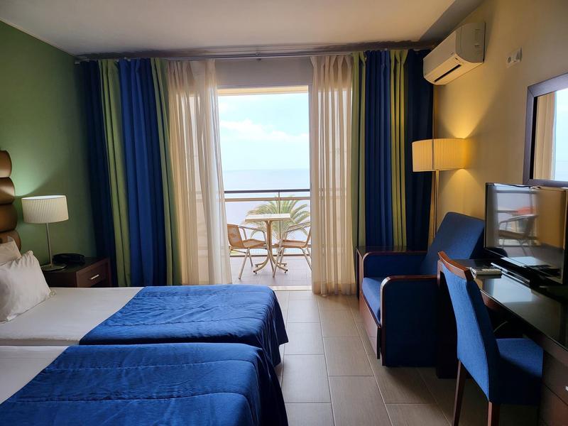 Pokój hotelowy z dwoma łóżkami, niebieskimi zasłonami, balkonem z widokiem na morze i miejscem do siedzenia.