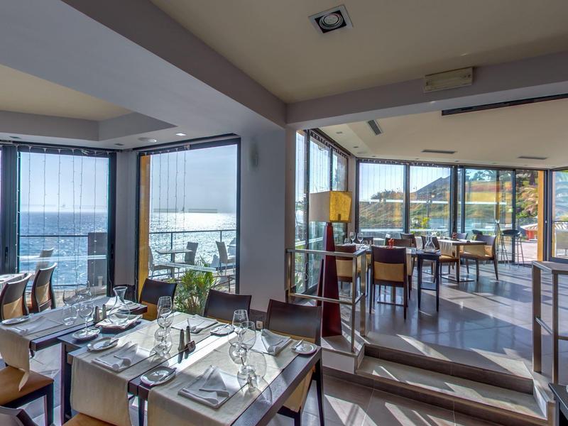 Jasna restauracja z stołami, krzesłami i panoramicznymi oknami z widokiem na morze.