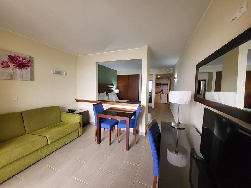 Nowoczesny pokój hotelowy z zieloną sofą, stołem do jadalni i dużym lustrem na ścianie.