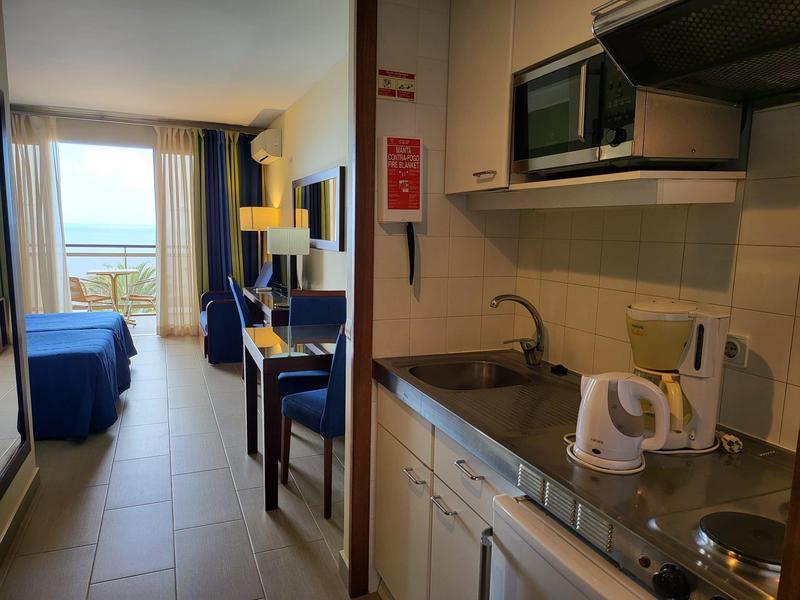 Pokój hotelowy z aneksem kuchennym, stołem jadalnym, częścią wypoczynkową i widokiem na morze przez duże okno.