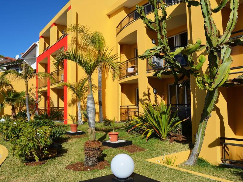 Żółty budynek hotelu z czerwonymi balkonami i tropikalnymi roślinami w ogrodzie w świetle słonecznym.