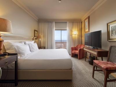 Chambre d'hôtel élégante avec lit double, fauteuils, télévision et vue sur le balcon.