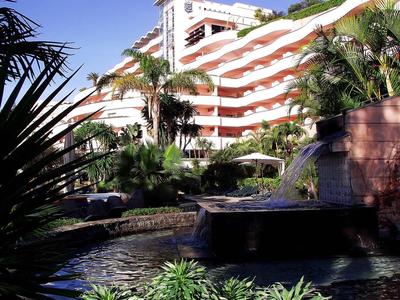Immeuble d'hôtel à plusieurs étages entouré de palmiers et d'un bassin d'eau sous un ciel bleu clair.