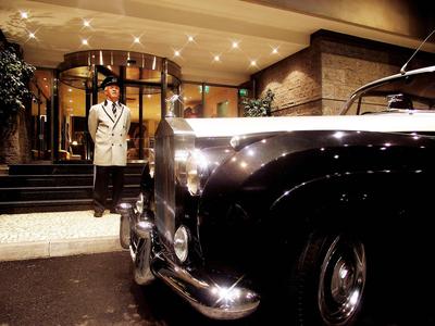 Een nette hotelportier staat naast een klassieke zwarte auto bij een ingang.
