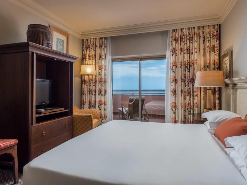Chambre d'hôtel confortable avec grand lit, TV, chaise et balcon avec vue sur la mer.
