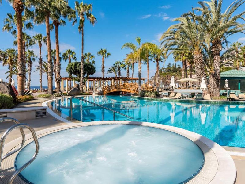 Piscine d'hôtel avec jacuzzi entourée de palmiers sous un ciel bleu clair.