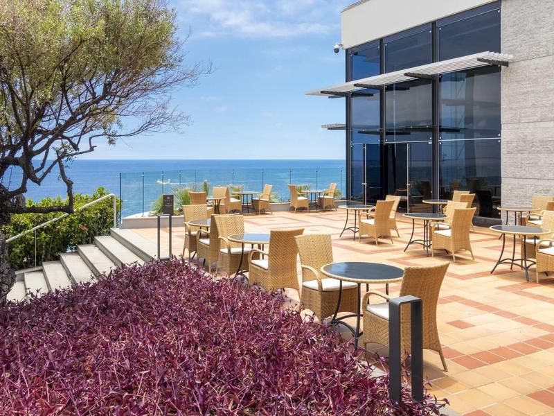 Terrasse avec tables et chaises devant un bâtiment moderne avec vue sur la mer.