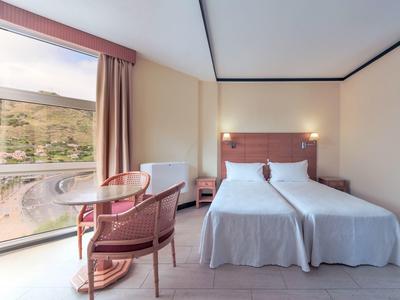 Světý hotelový pokoj se dvěma jednolůžky, skleněným oknem a výhledem na klikatou horskou cestu.