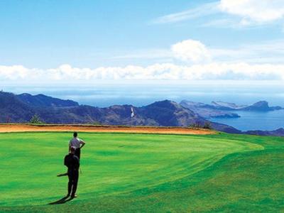 Golfista de pie en campo de golf verde con vista a montañas y mar bajo cielo despejado.