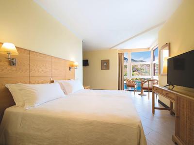 Světle zařízený hotelový pokoj s velkou postelí, televizí a balkonem s výhledem na hory.