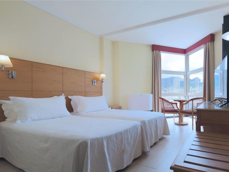 Světle zařízený hotelový pokoj se dvěma jednolůžky, posezením a velkým oknem s výhledem na hory.