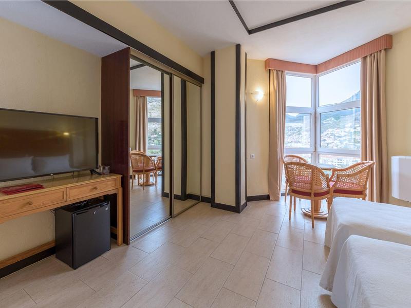 Světelný hotelový pokoj s manželskou postelí, televizí, psacím stolem a posezením u okna.