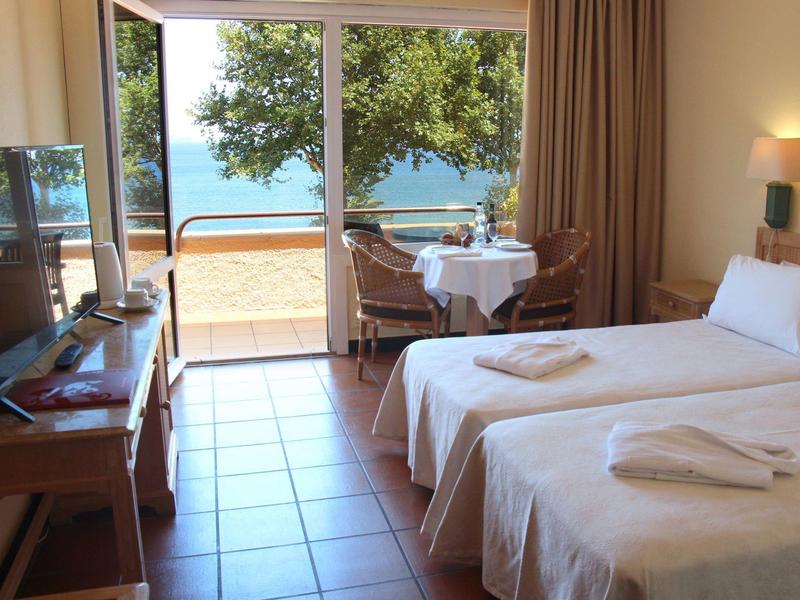 Světý hotelový pokoj se dvěma postelemi a výhledem na moře přes velké skleněné posuvné dveře.