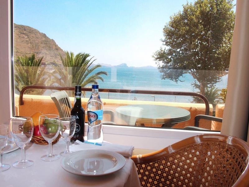 Pohled z hotelového pokoje na terasu s stolem, židlemi a mořem v pozadí.