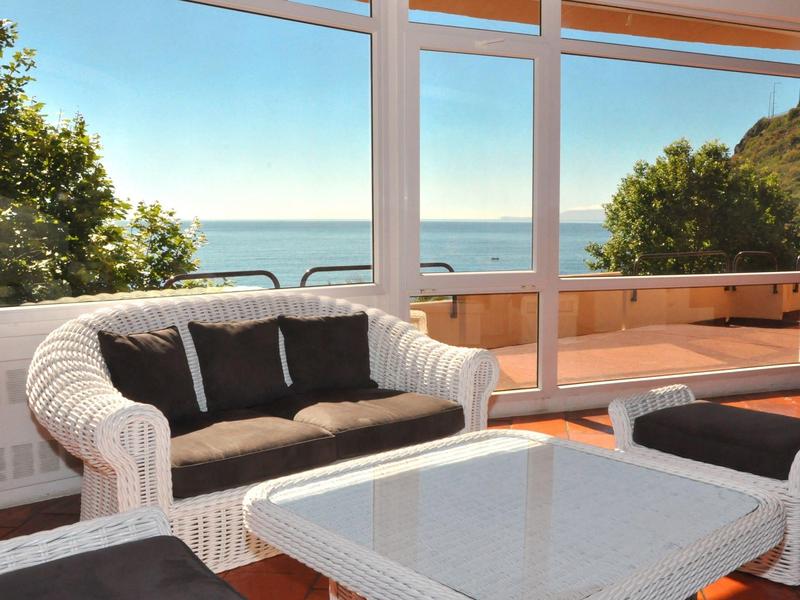 Pohodlné posezení s bílým ratanovým nábytkem a černými polštáři na slunné terase s výhledem na moře.