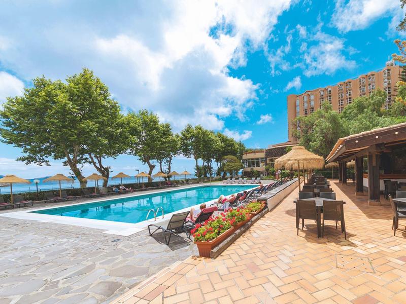 Venkovní bazén s lehátky a stolem vedle hotelu pod modrou oblohou s mraky.