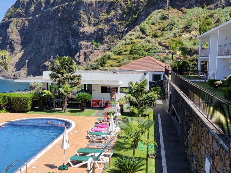 Hotel complex with pool, sun loungers, and palm trees against mountain backdrop.