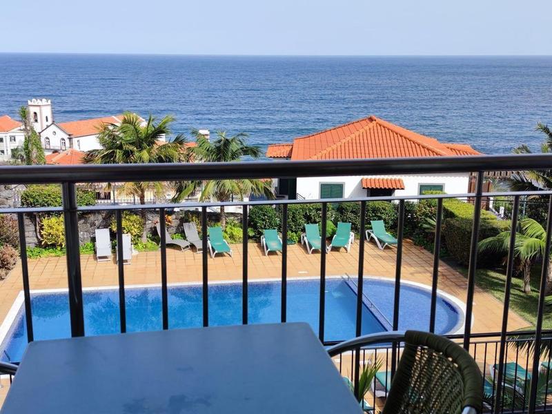 A balcony overlooking a pool surrounded by plants and buildings by the sea.