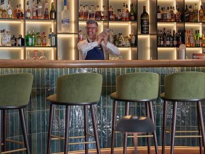 Bar con quattro sgabelli verdi e barista dietro il bancone illuminato