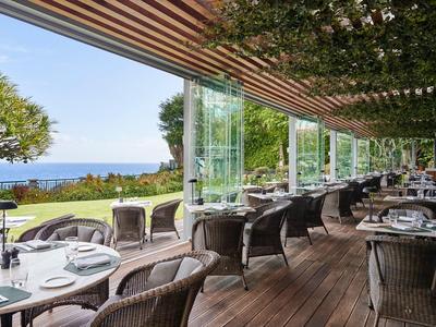 Terrazza dell'hotel con sedie in vimini e tavoli con vista su giardino e mare sotto un cielo limpido.