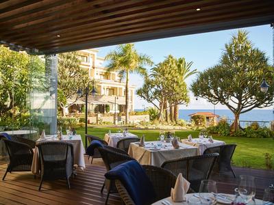Terrazza con tavoli apparecchiati con vista su giardino e mare, circondata da palme e alberi.
