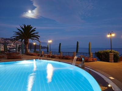 Piscina illuminata di notte con palme e mare sullo sfondo.